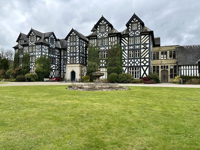 Large Tudor-style building with black and white timber facade, surrounded by gardens, lawn, and central stone fountain.