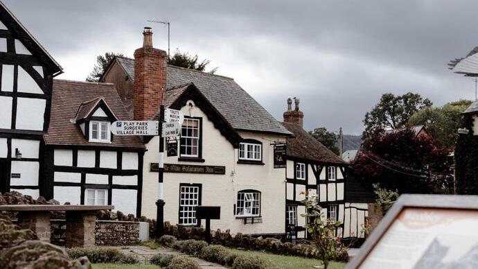 Historic black-and-white timber-framed buildings and an inn with stone walls and a garden under a cloudy sky.