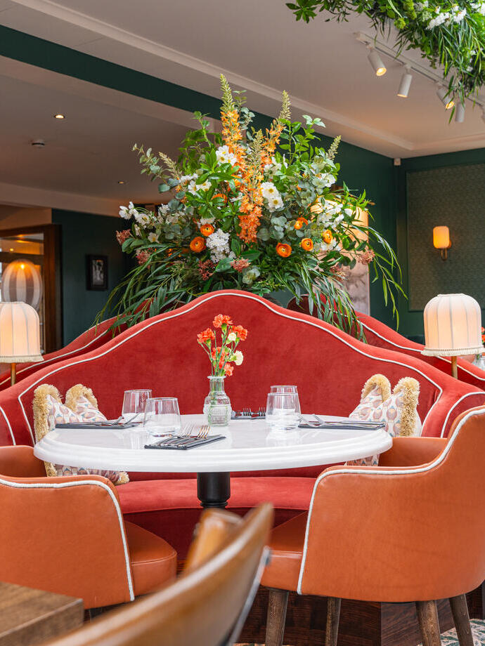 Stylish restaurant interior with orange chairs, a red velvet booth, floral arrangement, and greenery overhead.