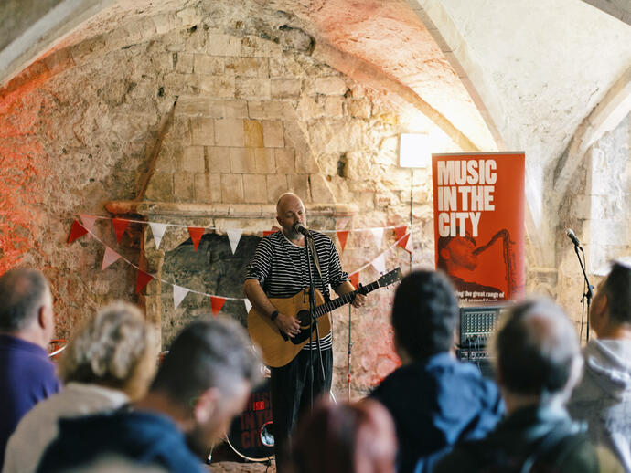 Musician performing with guitar in historic stone building, 'Music In The City' event banner and audience in foreground.