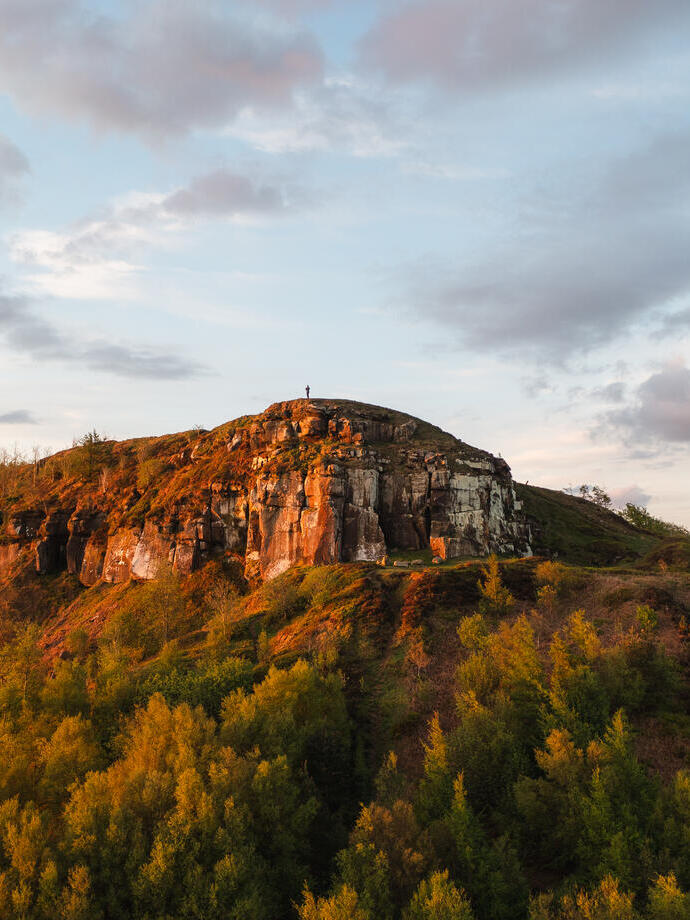 A lone person stands on a cliff top with forest below
