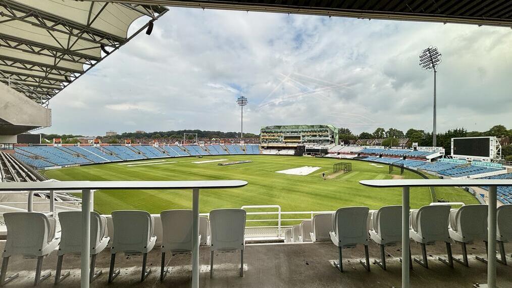 View from behind white seats and tables overlooking a large cricket stadium with green grass, blue stands, and floodlights under a cloudy sky.