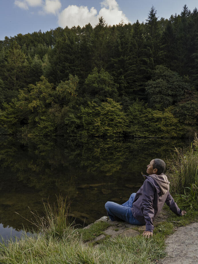 Person relaxing by a calm lake surrounded by dense green forest and clear sky, reflecting tranquility and nature.