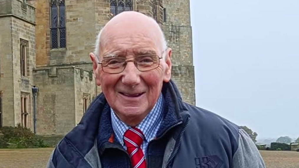 Smiling older man with glasses standing outside a historic stone building on a cloudy day.