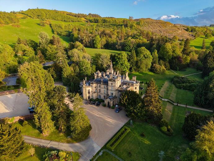 Aerial view of a grand historic manor surrounded by lush gardens, large trees, and rolling green hills under a blue sky.