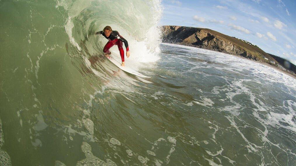 Man surfing a wave, crouched low on the board with cliffs in the distance