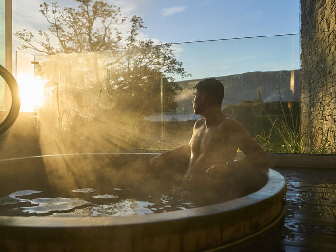 A man relaxes in a steaming outdoor hot tub as the sun rises, surrounded by nature and modern stone architecture.