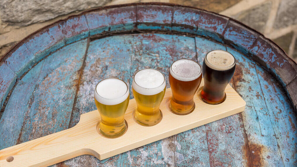 Quatre types de bière dans des petits verres sur une planche en bois, posés sur un tonneau bleu vieilli avec un mur en pierre en arrière-plan.