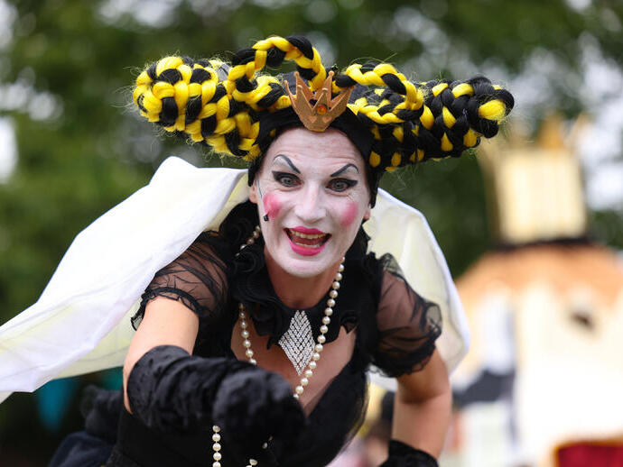 Person in elaborate costume and dramatic makeup with black and yellow braided hair outdoors, trees and event scenery in the background.