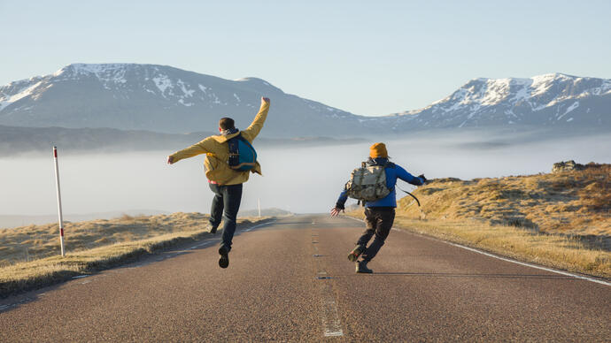 Two men walking and jumping on a road surrounded by mountains