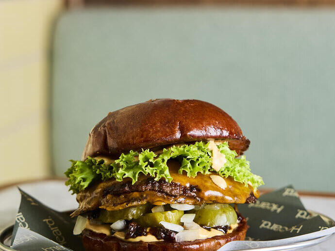 Close-up of a gourmet burger with lettuce, cheese, pickles, onions, and sauce in a glossy brioche bun on a plate in a restaurant setting.