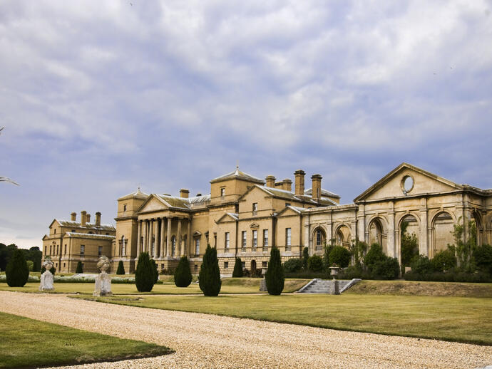 Large neoclassical mansion set in landscaped gardens with a gravel path, manicured lawns, and overcast sky.