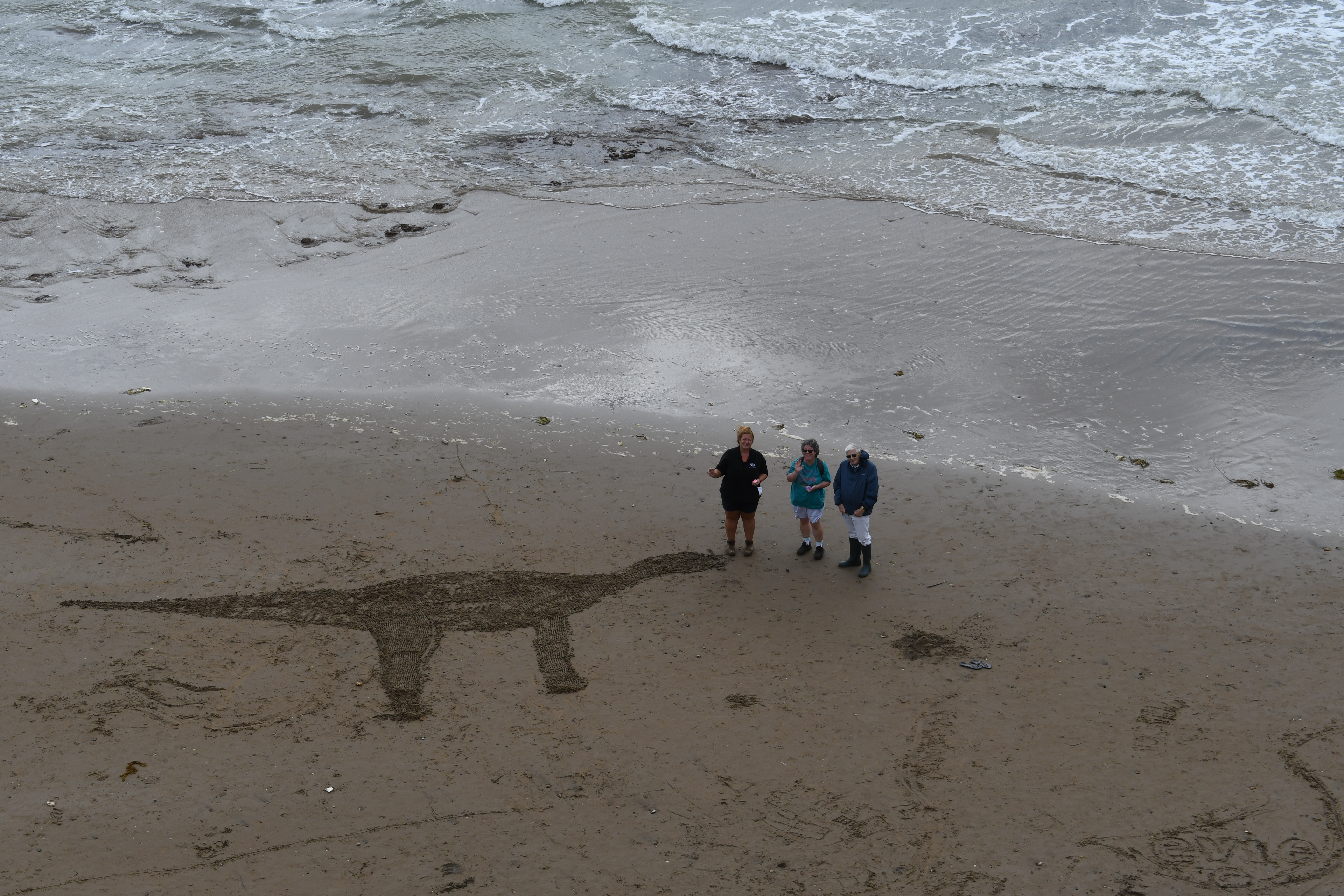 People on a beach with a dinosaur carved into the sand