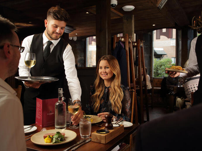 A smartly dressed staff member serving wine and food to a couple in a dining room 