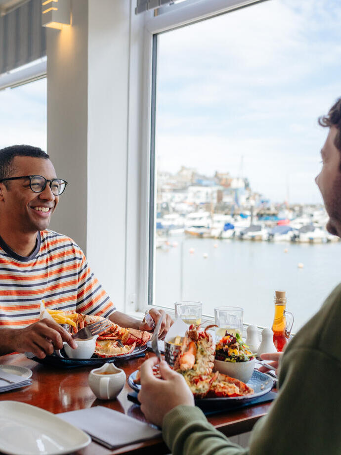 Two men eat lobster in a restaurant with a harbour view