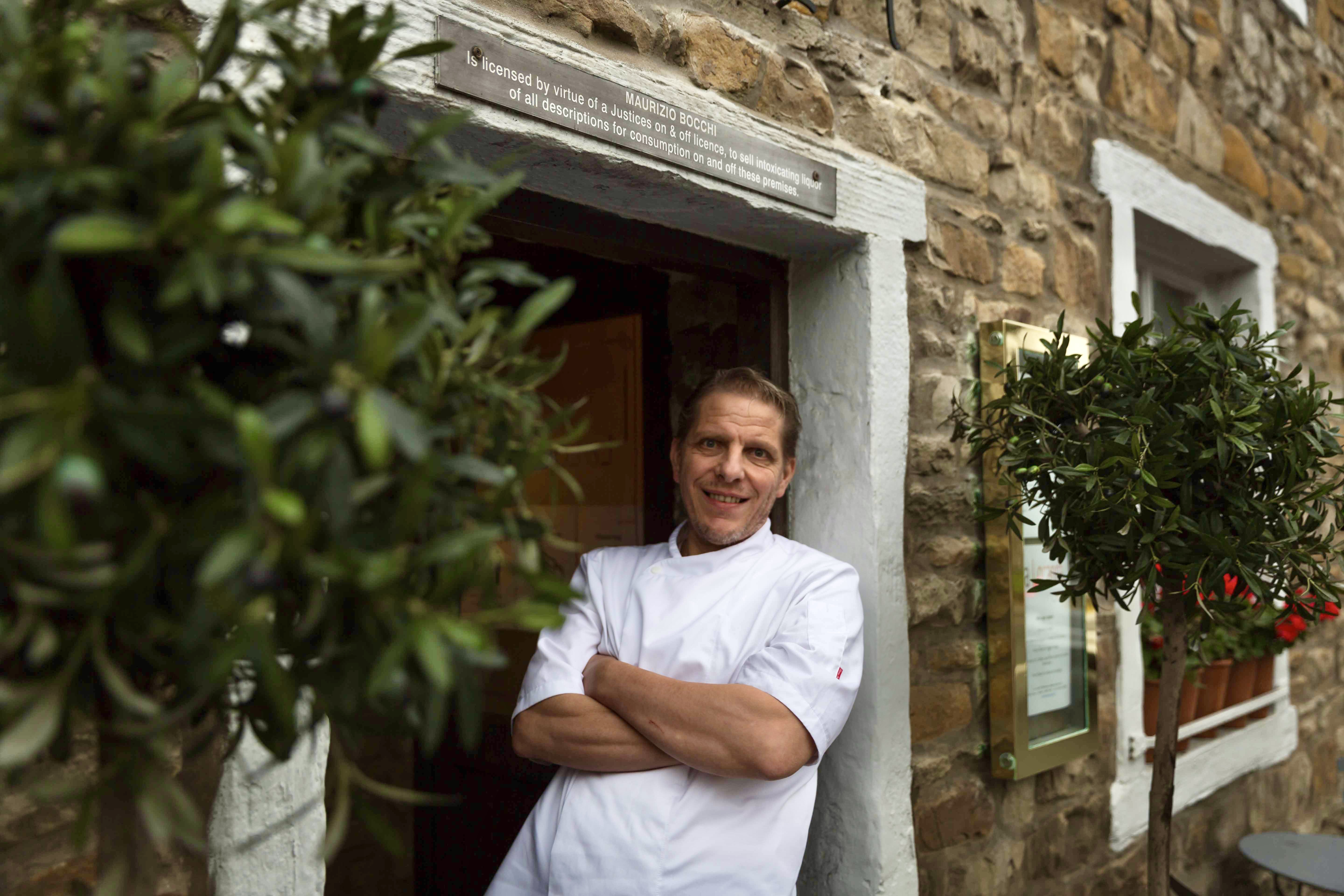 Chef in white uniform stands smiling at the entrance of a rustic stone building with white window frames and potted plants.