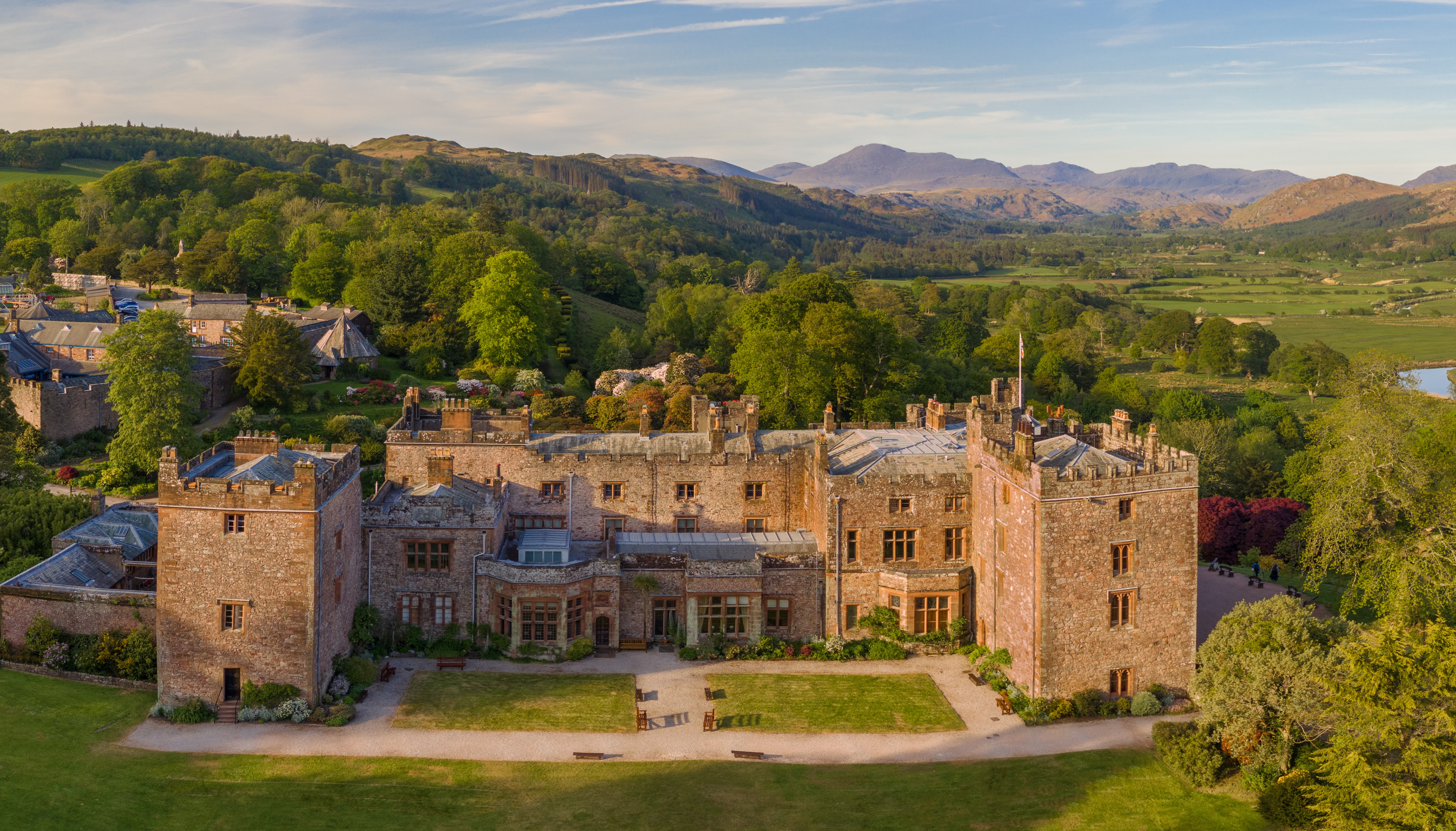 A country house with decorative mock foundations against a backdrop of tree covered hills, including Scafell Pike. Muncaster Castle - Silver award winner for Small Visitor Attraction of the Year at the VisitEngland Awards for Excellence 2023.