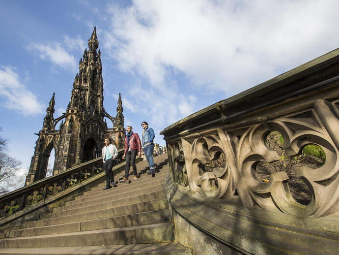 Three people walking down stone steps near the ornate Scott Monument under a partly cloudy blue sky in Edinburgh.