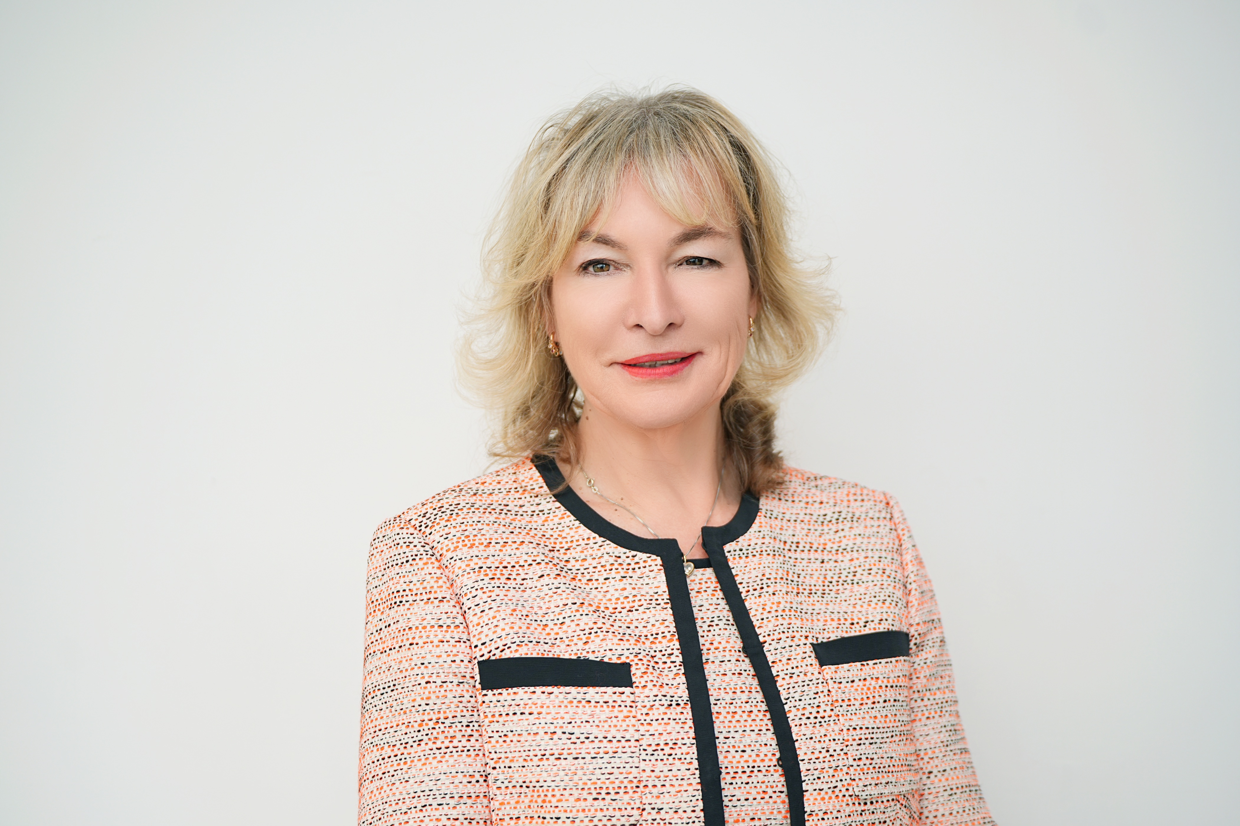 A woman with blonde hair wearing a light patterned jacket with black trim, standing against a plain white background.