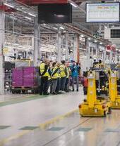 People in safety vests tour a modern factory with machinery, assembly lines, and overhead digital displays.