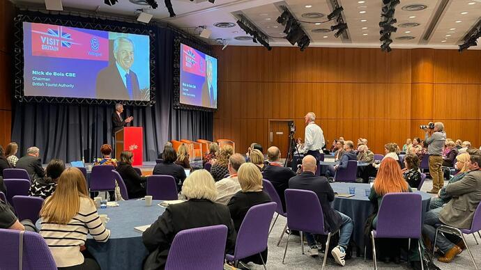 BTA Chairman Nick de Bois CBE stands behind a podium onstage and speaks to a room of 80 delegates at the VisitBritain Business Events Association Conference