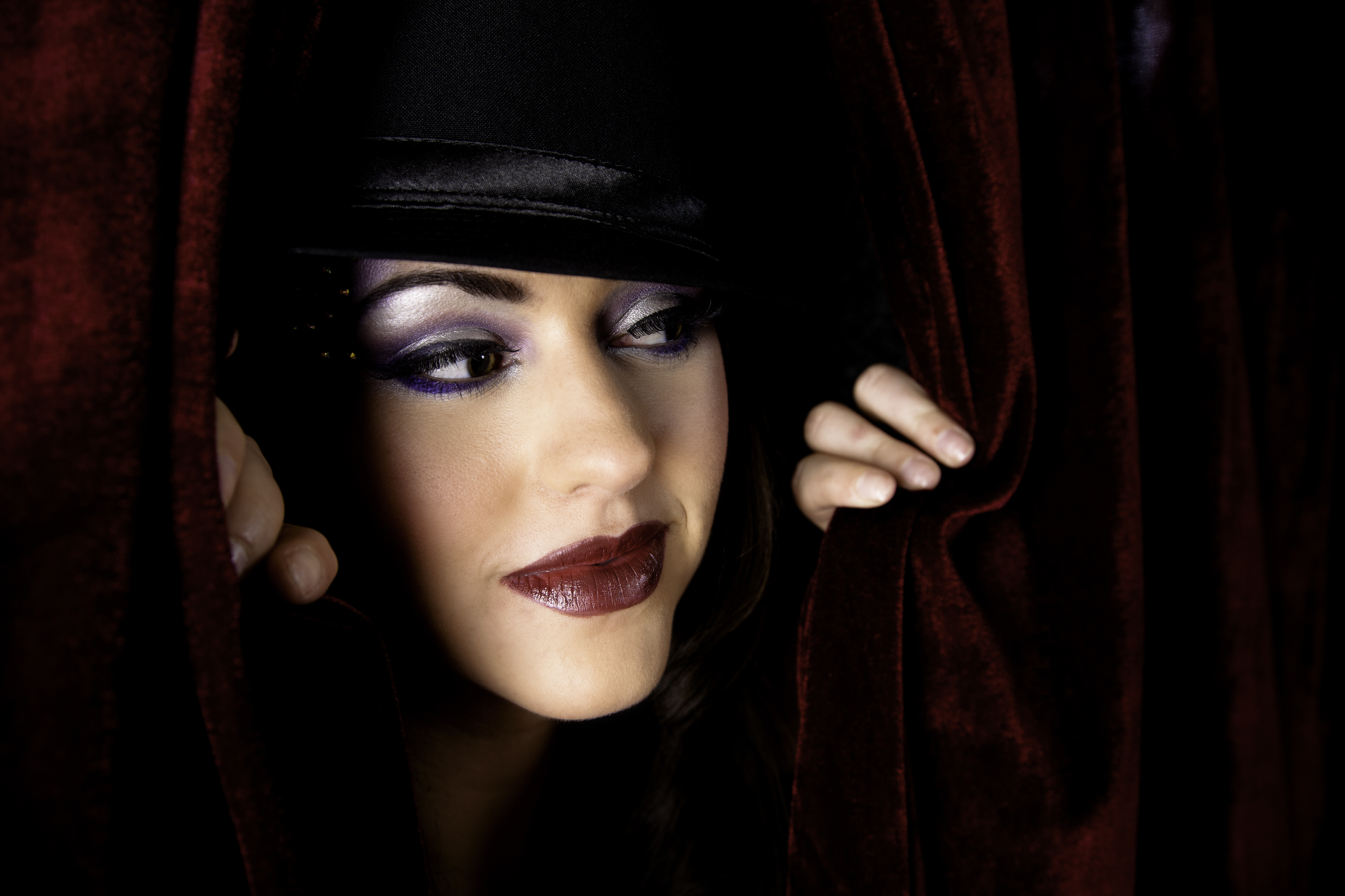 A performer wearing a hat and in full make up peaking out from behind a curtain