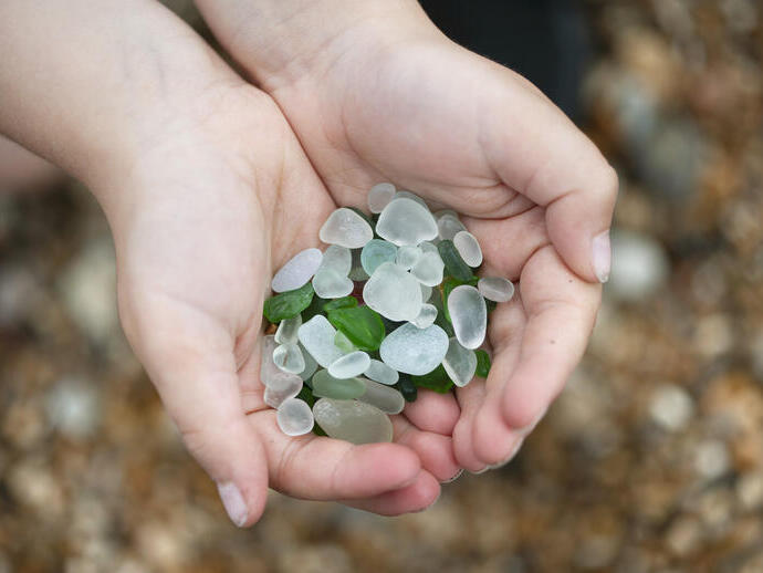 Hands holding pieces of white and green sea glass, with a blurred background of pebbles or sand.
