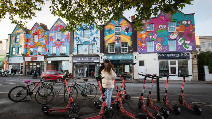 Zwei Personen stehen neben Leihrollern vor einer Reihe von Häusern, die mit bunten Wandmalereien verziert sind