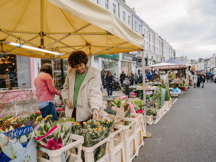 Outdoor flower market scene on a city street with people shopping at stalls under yellow tents, surrounded by historic buildings.