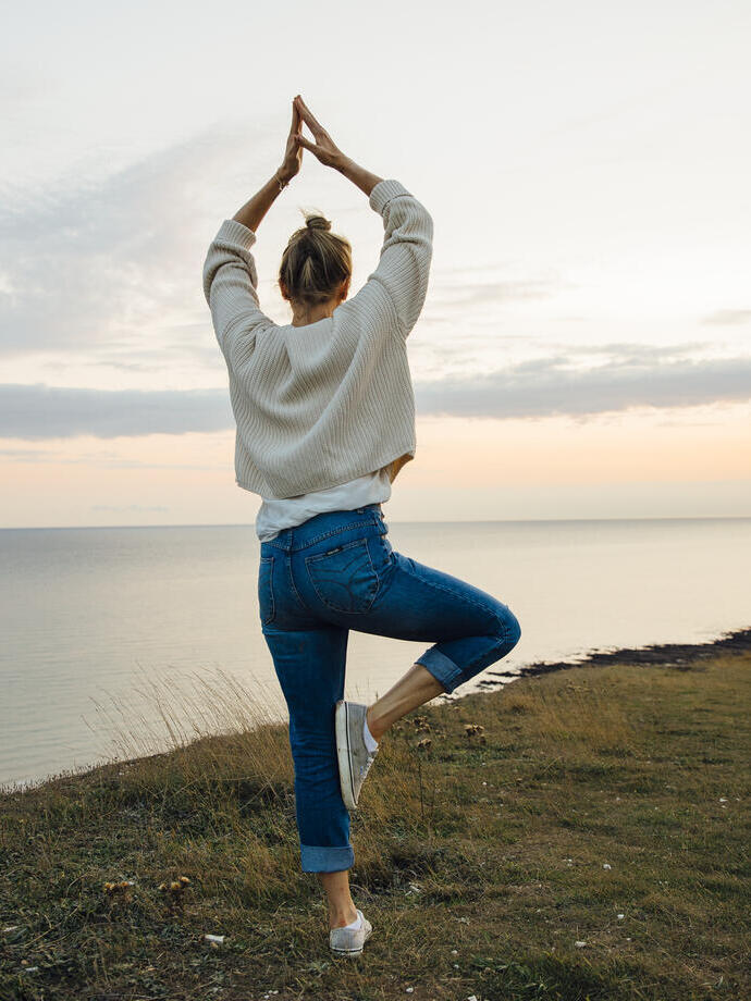 Frau in einer Yoga-Pose, die auf einer Klippe steht und auf das Meer blickt