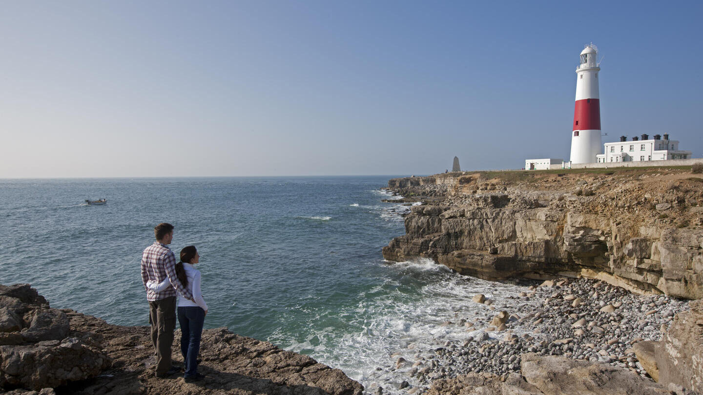 Paar steht auf einer Klippe am Meer und schaut auf einen rot-weißen Leuchtturm