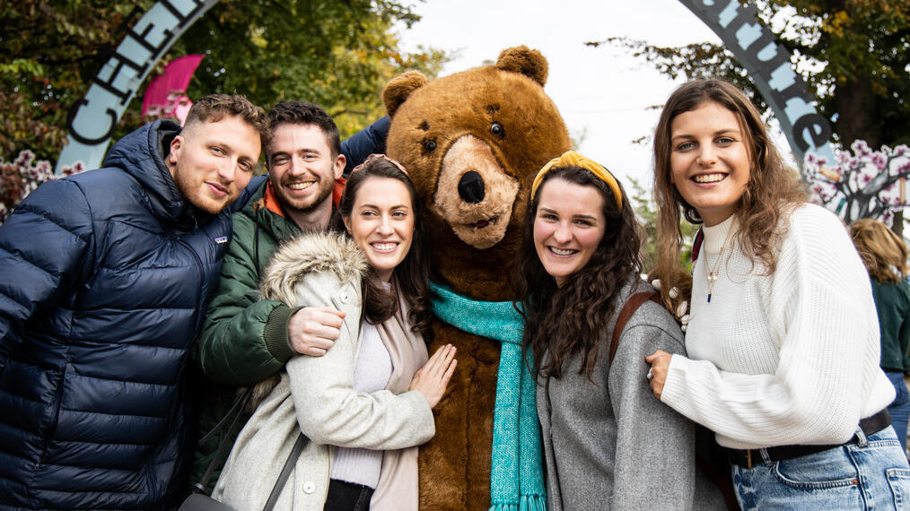 Gente posando con una gran mascota en forma de oso bajo un arco con el letrero «Cheltenham Literature»