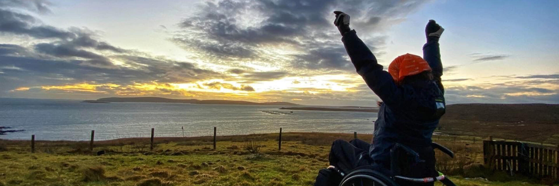 Une personne en fauteuil roulant, les bras tendus, regardant la mer et admirant le lever du soleil