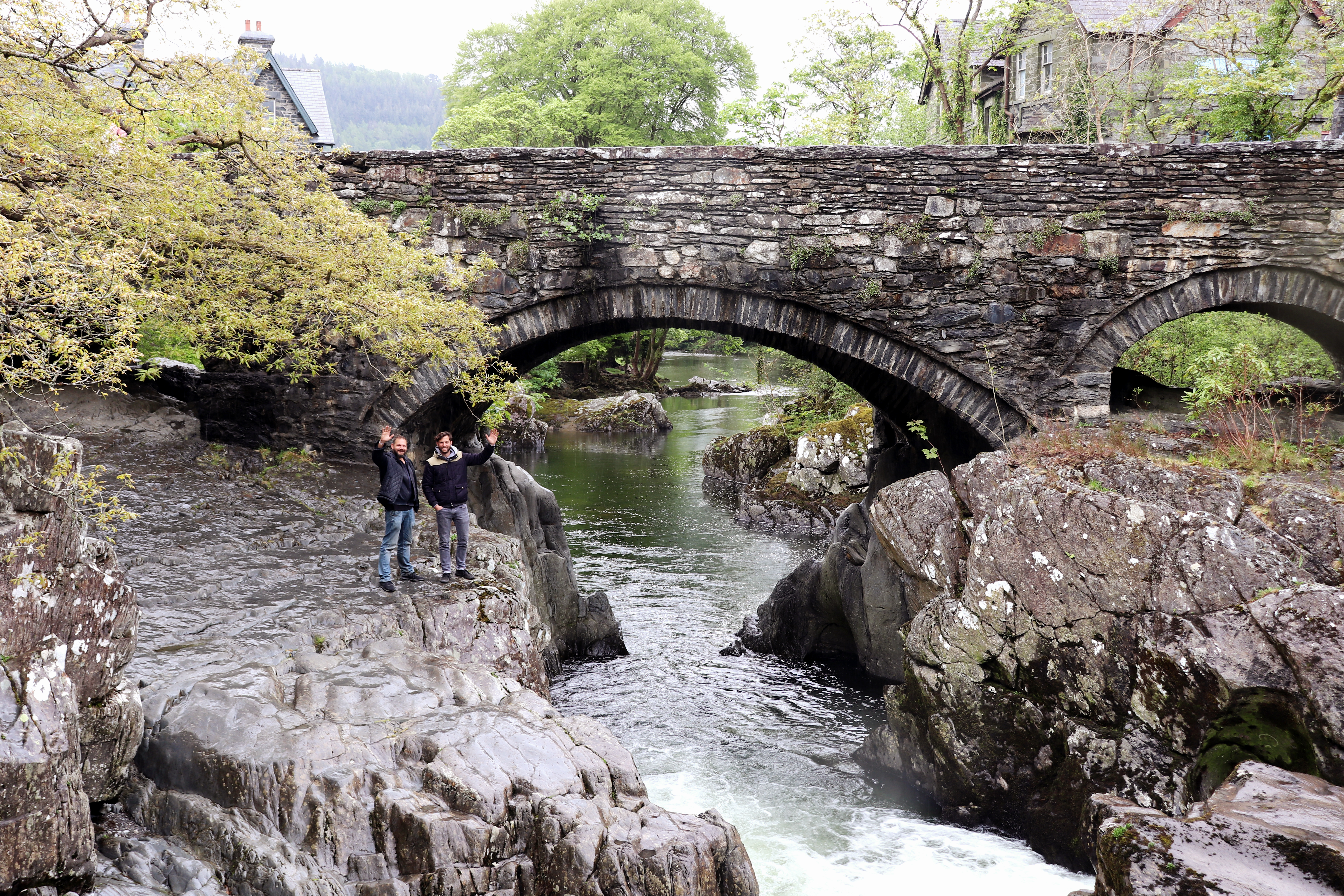 Two men waving from some rugged rocks beneath a stone bridge over a narrow river