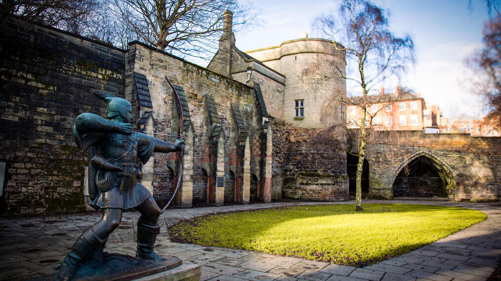 Statue of an archer in front of historic stone castle walls, green lawn, trees, and blue sky in the background.