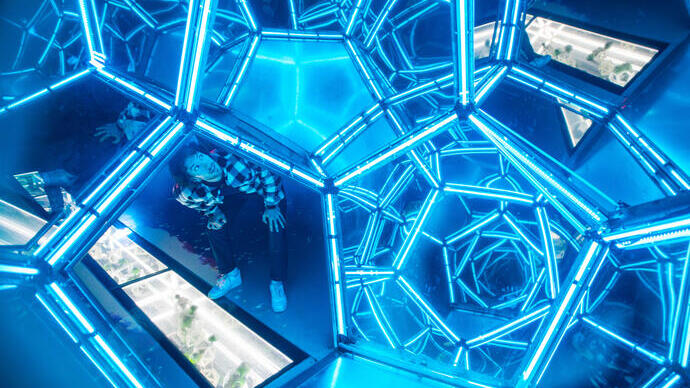 Person inside a geometric room with blue neon lights and reflective surfaces creating a kaleidoscopic effect.