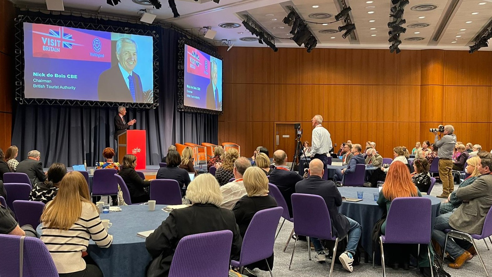 BTA Chairman Nick de Bois CBE stands behind a podium onstage and speaks to a room of 80 delegates at the VisitBritain Business Events Association Conference