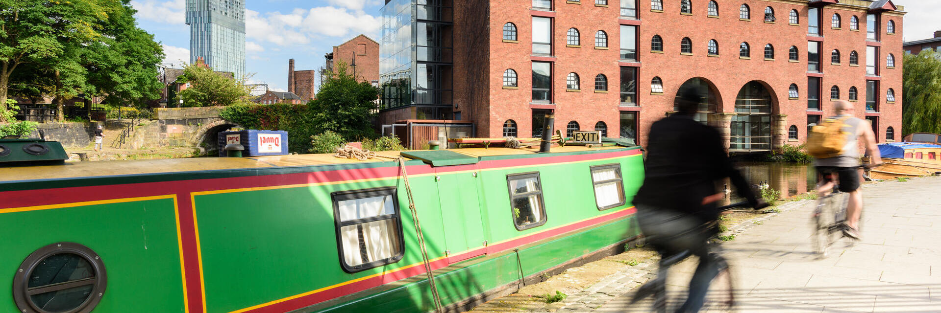 Canal boat moored by waterside, red brick warehouse, modern skyscraper, cyclists passing, sunny urban scene.