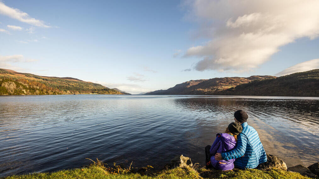 Two people in jackets sit by a calm lake, surrounded by hills and trees under a blue sky with scattered clouds.