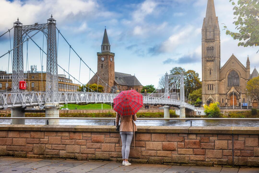 A woman with an umbrella facing a river next to a bridge towards a church