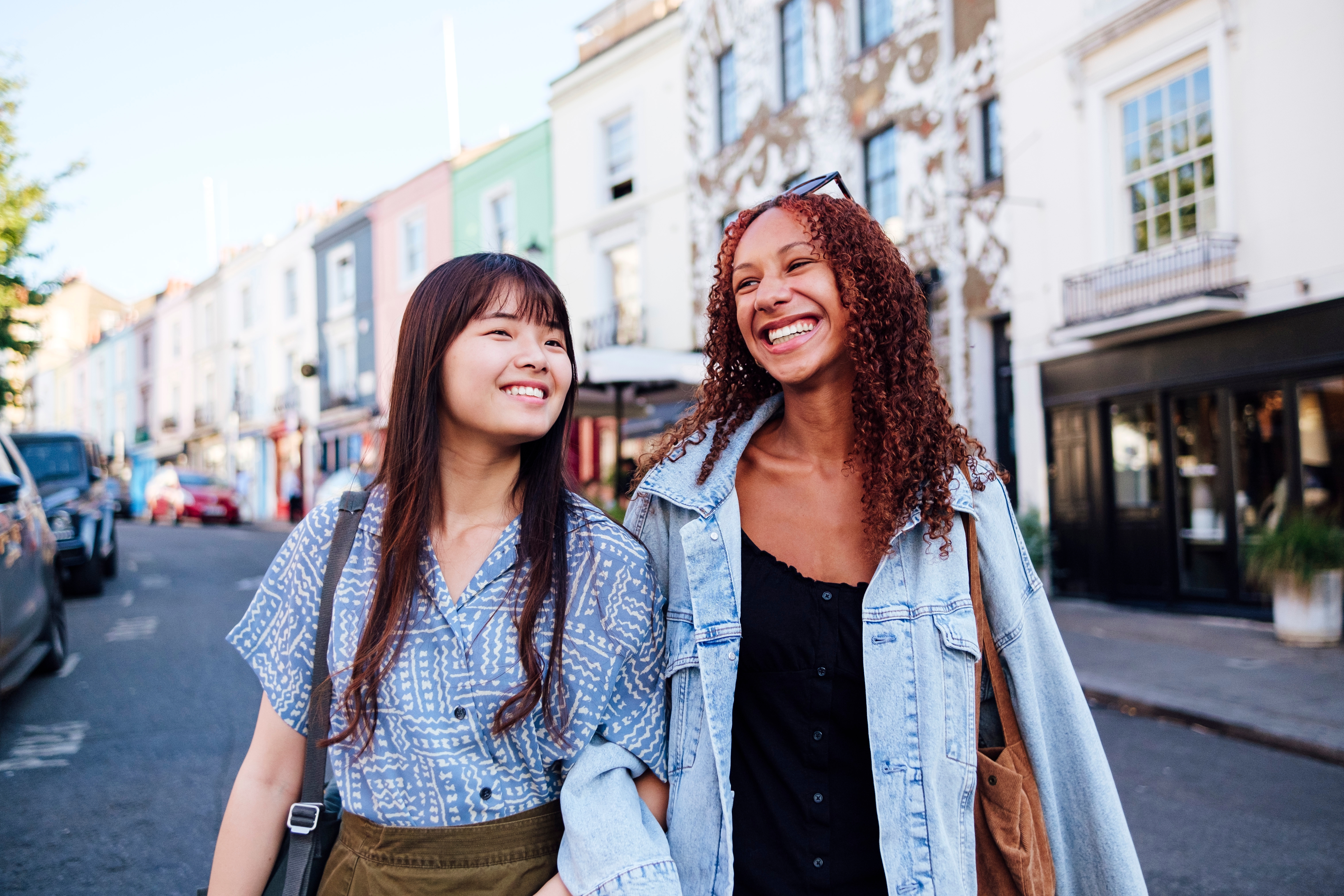 Two young women linking arms and walking in a street smiling