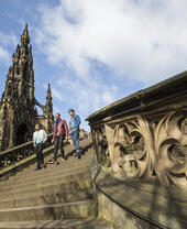 Three people walking down stone steps near the ornate Scott Monument under a partly cloudy blue sky in Edinburgh.