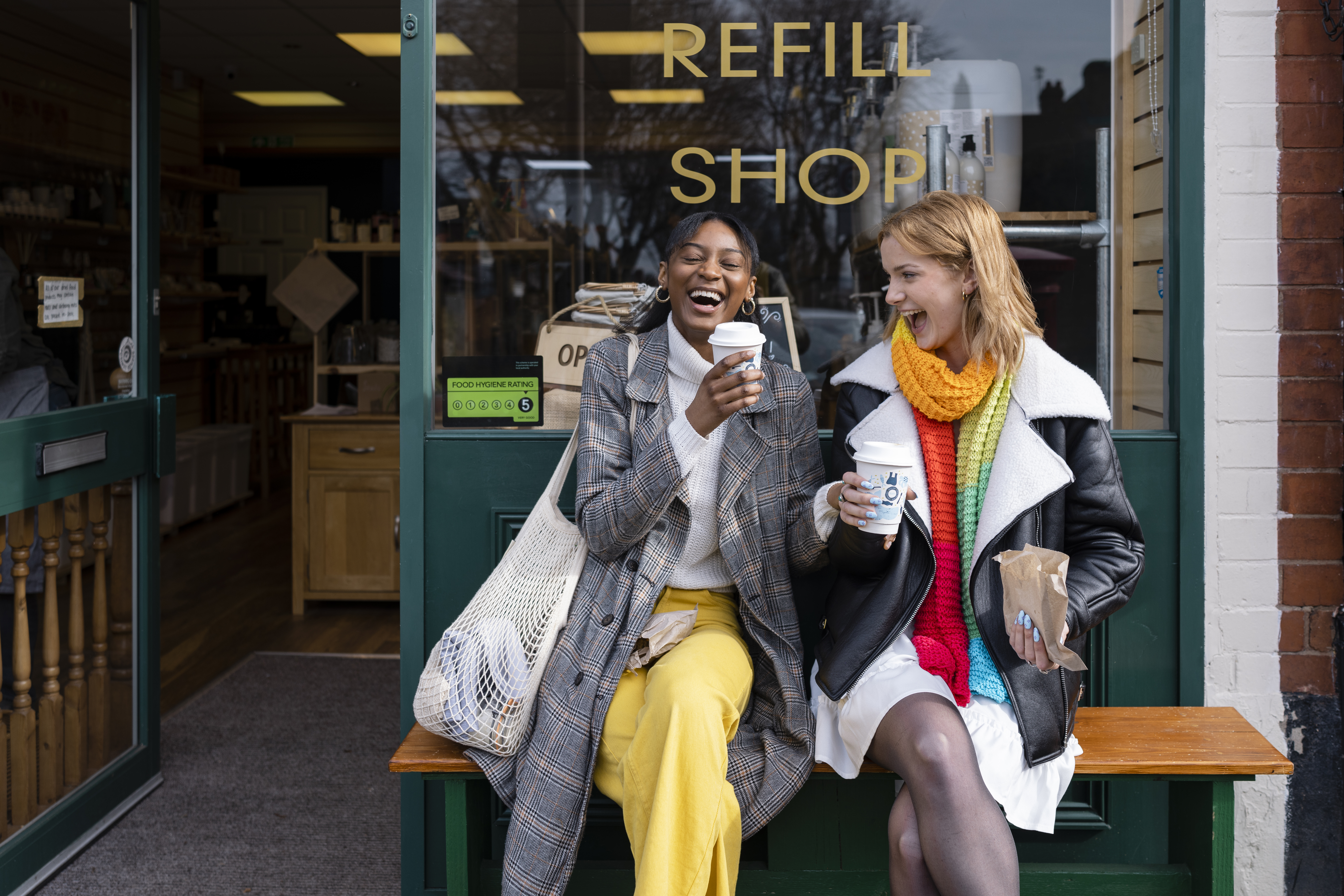 Two young women sat on a bench outside a coffee shop with takeaway coffees