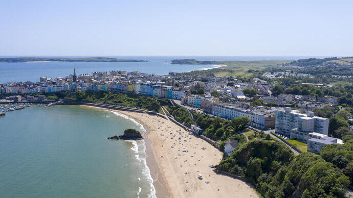 An overhead shot of a sandy beach lined with sunbathers, surrounded by brightly coloured townhouses and countryside.
