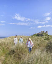 A group of people walk among tall grasses in dunes with a castle in the background
