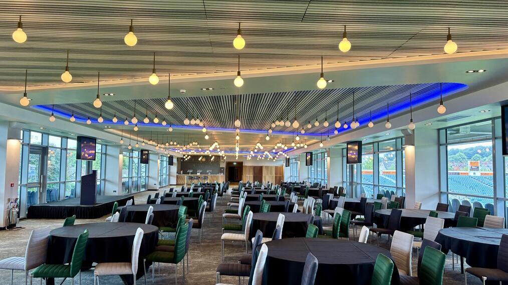Spacious modern event space with round tables, colorful chairs, large windows, and decorative ceiling lights overlooking a sports stadium.
