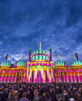 Crowd sitting on the ground in front of a large ornate building lit up with bright colours