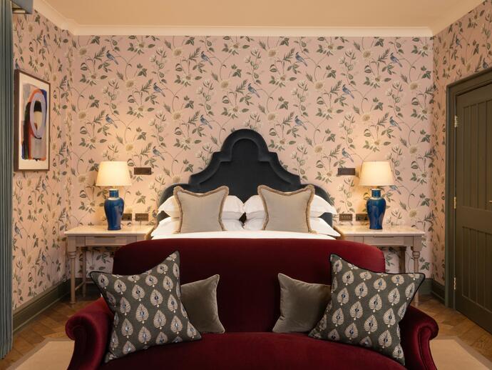 Luxuriöses Hotelzimmer mit rotem Samtsofa am Fußende des Bettes und blumiger Tapete an den Wänden dahinter.