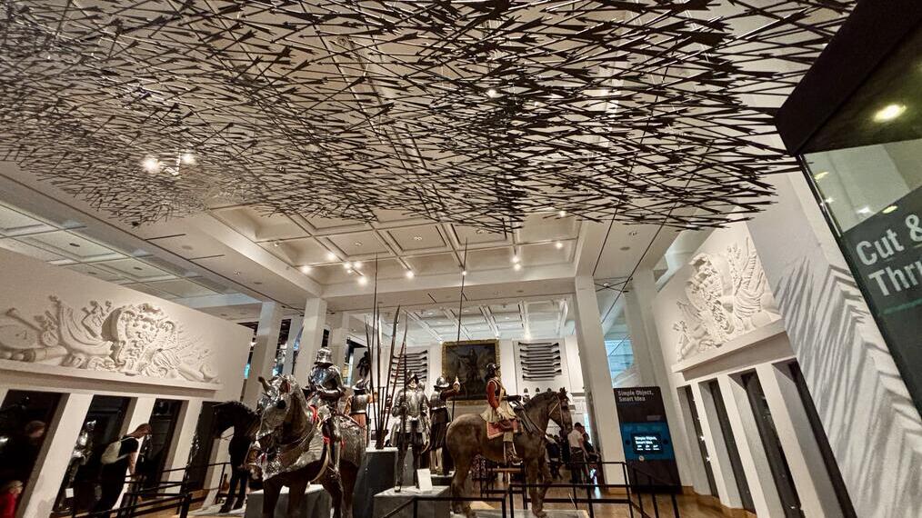 Exhibit of armored knights on horseback with weapons, intricate ceiling display, and museum visitors observing the scene.