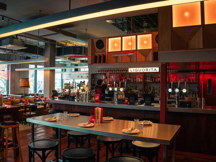 Modern restaurant interior with wooden decor, bar area, stools, tables set for dining, and warm lighting.